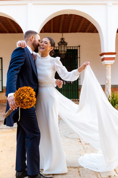 Pareja sonriente en un entorno romántico, vestidos de boda, sosteniendo un ramo de flores.